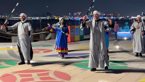 Arabische-Harmonie-Tanzende-Männer-Und-Frauen-Zusammen-In-Doha,-Katar.-Gruppenauftritt-Bei-Einer-Hellen,-Fröhlichen-Partynacht.-Lokaler-Traditioneller-Tanz-Mit-Musik-Am-Strand-Von-Corniche.-Das-Meer-Spiegelt-Die-Stadtlandschaft-Wider.