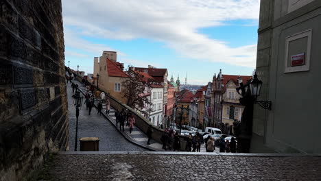 Toma-Del-Colorido-Encanto-De-La-Calle-Nerudova-Y-Las-Históricas-Escaleras-Del-Castillo-De-Praga,-Con-Gente-Paseando-Tranquilamente-Desde-El-Castillo-Y-Postes-De-Luz-Clásicos-Bordeando-La-Calle.