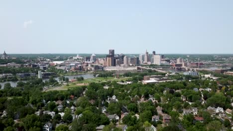 Wide-aerial-shot-slowly-moving-forward-with-the-city-of-Saint-Paul,-Minnesota-on-the-horizon-next-to-the-Mississippi-river-surrounded-by-trees-and-houses-on-a-sunny-day