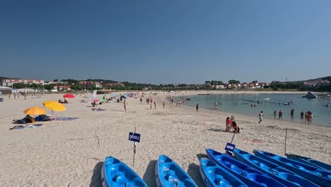 people-enjoying-a-morning-on-the-beach-of-crystal-clear-water,-sunbathing,-swimming,-walking,-canoeing,-european-holiday-town,-sunny-morning,-descriptive-panning