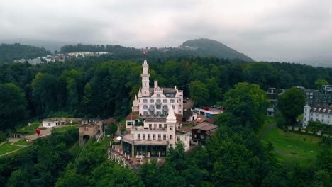 Aerial-view-towards-the-Chateau-Gutsch-castle,-restaurant-and-hotel,-on-a-hill-in-Luzern-city,-dark,-moody,-summer-day,-in-Lucerne,-Switzerland---tilt-down,-drone-shot