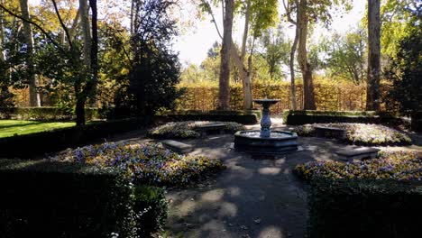 Toma-Panorámica-De-Una-Pequeña-Fuente-Rodeada-De-Bancos-De-Piedra-En-El-Parque-Del-Capricho-En-Madrid,-España.