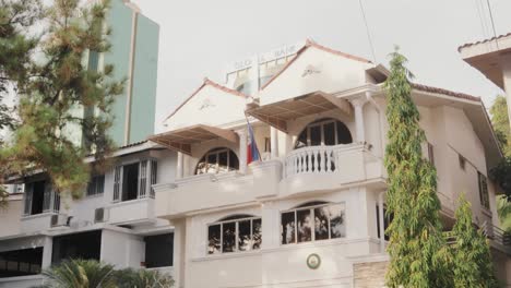 Haitian-Ambassy,-Architectural-beautiful-white-house-with-three-floors,-surrounded-by-trees-and-has-a-skyscraper-in-the-background