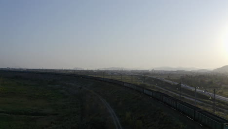 Full-loaded-coal-cargo-freight-train-locomotive-stationary-along-a-high-rise-railway,-in-green-fields-with-mountain-ridge-in-the-far-distance,-on-the-sunset