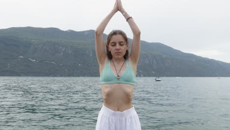 Young-Woman-lifts-her-arms-Yoga-pose-hands-together-below-mountain-lake-panorama