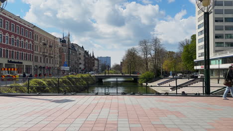 A-man-walks-past-Davids-Halls-Bridge-in-Malmö,-with-the-Kronprinsen-shopping-center-and-the-'Way-to-Go'-artwork-in-the-background