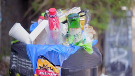 Overfilled-public-litter-bin,-trash-can-filled-with-plastic-bags-and-glass-bottle,-by-city-canal-with-jogger-in-the-background