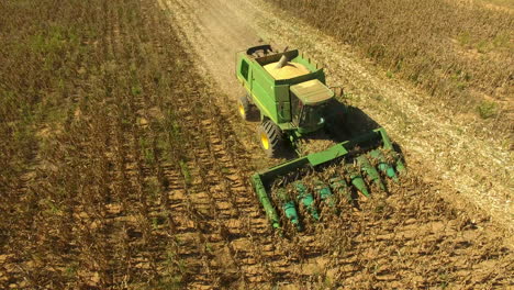 Wide-aerial-shot-of-the-front-of-a-John-Deere-combine-harvester-tractor-with-spinning-corn-heads-moving-in-a-straight-line-across-a-golden-field-that-pans-up-to-see-the-blue-sky