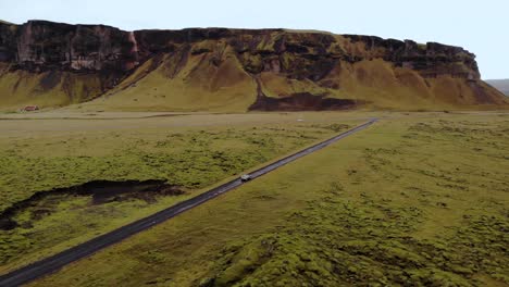 Aerial-4k-view-of-a-SUV-car-driving-on-a-gravel-road-surrounded-by-green-moss-lava-field-with-a-beautiful-mountain-landscape-in-Iceland,-Europe,-orbit-shot