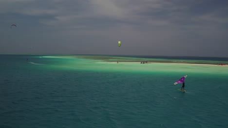 Colorful-kitesurfers-glide-over-clear-blue-waters-near-a-sandy-island-in-the-bright-daylight