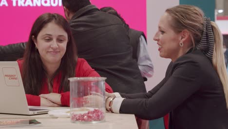 Two-professional-women,-seated-and-talking-to-each-other,-with-laptop-at-FITUR,-Spain