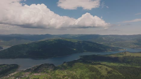 Aerial-pan-shows-an-accumulation-lake-surrounded-by-green,-forested-mountains,-partly-cloudy