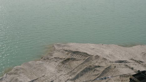 Aerial-flyby-shot-of-abandoned-heavy-machines-on-the-bank-of-a-lake-on-a-bright-day