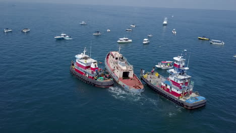 Aerial,-ship-being-prepared-to-be-sunk-in-ocean,-surrounded-by-small-boats