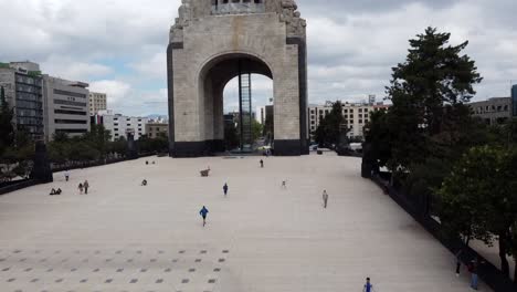 Vista-Aérea-De-Personas-Corriendo-Hacia-Un-Monumento-En-Un-Día-Nublado-En-La-Ciudad-De-México