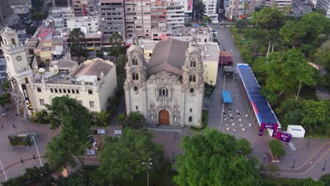 Drone-orbits-around-church-called-"Virgen-Milagrosa"-surrounded-by-green-trees-of-park-called-"Parque-Kennedy"-Located-in-Miraflores-district-of-Lima,-Peru