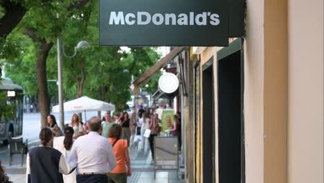 Pedestrians-walk-past-the-American-multinational-fast-food-hamburger-restaurant-chain,-McDonald's,-and-its-street-sign-in-Madrid,-Spain