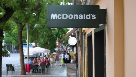 People-walk-past-the-American-multinational-fast-food-hamburger-restaurant-chain,-McDonald's,-and-its-street-sign-in-Madrid,-Spain