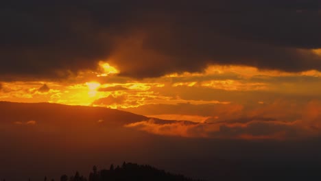 Aerial-shot-rising-above-silhouetted-mountain-ridge-revealing-orange-sunset-as-it-casts-a-warm-glow-between-a-cloudy-sky-and-distant-mountain-ranges,-high-contrast