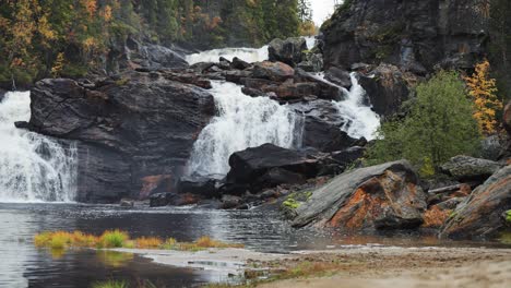 Im-Norwegischen-Herbstwald-Stürzt-Ein-Majestätischer-Wasserfall-über-Dunkle-Felsen-Und-Bietet-Blick-Auf-Einen-Ruhigen-See-Und-Einen-Sandstrand-Darunter,-Wodurch-Eine-Bezaubernde-Szenerie-Natürlicher-Schönheit-Entsteht.