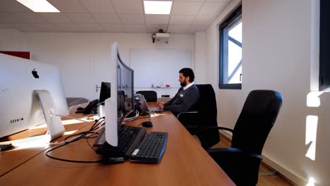 Toma-De-Seguimiento-De-Una-Sala-De-Trabajo-A-Nivel-De-Oficina,-Dos-Hombres-Trabajando-En-Computadoras,-Pantallas-Grandes-En-Primer-Plano,-Luz-Del-Día