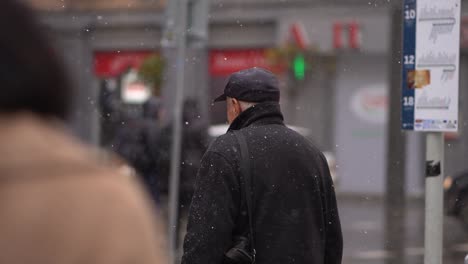 Old-European-man-looks-around-crowded-street,-snow-falls-slow-motion