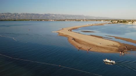 Aerial-panorama-of-a-Nin-town,-sandy-beach-and-boat-approaching-in-sunset-with-mountain-range-Velebit-in-the-distance