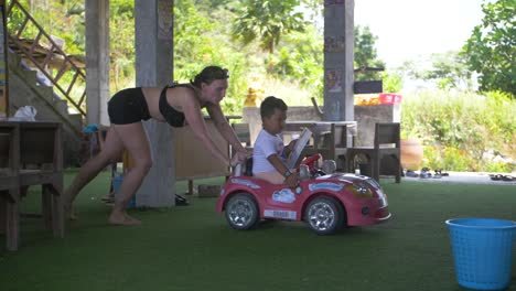 Woman-in-black-shorts-pushing-a-small-pink-toy-car-with-little-boy-riding-inside