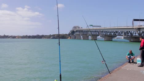 Dos-Hombres-Haciendo-Preparativos-Para-La-Pesca-En-Westhaven,-Auckland,-Nueva-Zelanda,-El-Puente-Del-Puerto-En-Segundo-Plano.