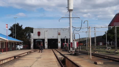 Two-people-walk-through-old-train-yard-with-train-workshop-in-background,-wide-shot