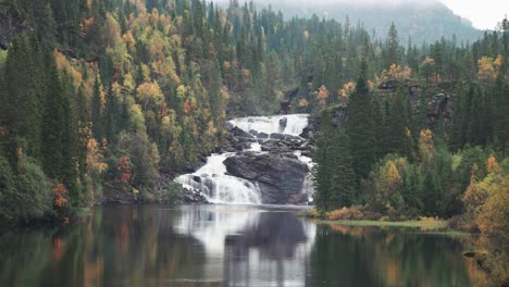 Amidst-the-Norwegian-autumn-forest,-a-waterfall-cascades-over-dark,-withered-cliffs,-its-beauty-mirrored-on-the-tranquil-surface-of-a-nearby-lake