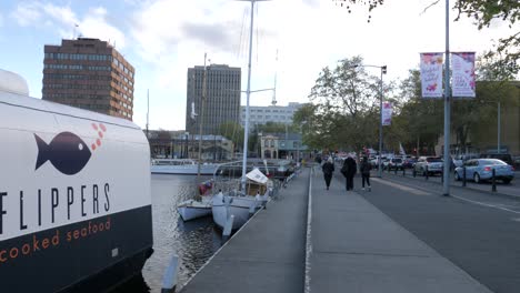 Gente-Caminando-Por-El-Paseo-Marítimo-De-Hobart-Con-Una-Batea-De-Pescado-Local-En-Primer-Plano,-Hobart,-Tasmania