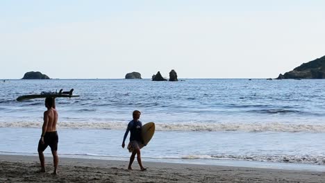 Dos-Jóvenes-Surfistas-Llevando-Sus-Tablas-De-Surf-A-Lo-Largo-De-Playa-Playitas-Cerca-De-Manuel-Antonio,-Costa-Rica-Durante-La-Puesta-De-Sol