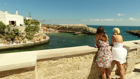 Dos-Mujeres-Jóvenes-Disfrutando-De-Una-Vista-Romántica-De-La-Playa-De-Polignano-A-Mare-Cala-Paura-Desde-Una-Terraza-En-Un-Día-Ventoso-De-Verano