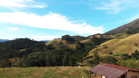 Aerial-view-of-a-small-rural-house-with-a-fenced-yard-in-a-hilly-landscape,-under-a-bright-blue-sky