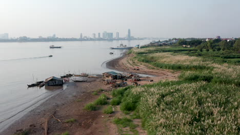 Aerial-drone-footage-flying-over-a-fisherman-floating-house-on-the-Mekong-shore-and-revealing-Phnom-Penh-on-the-background