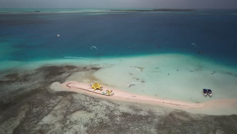 Kite-surfers-gliding-near-a-small-sandy-island-with-a-colorful-tent-and-clear-waters,-aerial-view