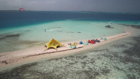 Colorful-kitesurfing-tents-on-a-sandy-beach-island-surrounded-by-turquoise-waters,-aerial-view