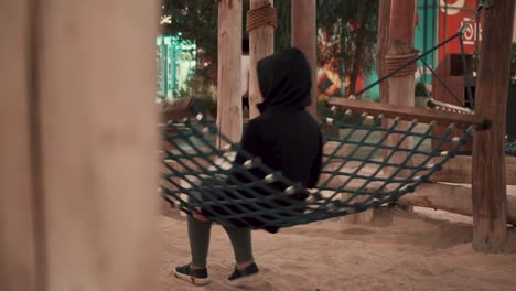 A-mysterious-and-strange-pan-view-over-a-pillar-of-a-lady-back-in-black-hoody-and-shoes-swinging-on-a-net-hammock-in-a-park,-A-slow-mo-view