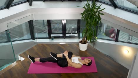 Woman-practicing-pilates-knee-to-chest-pose-on-mat-in-bright-indoor-space,-modern-architecture