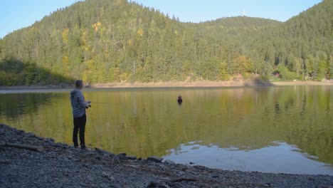 Young-man-is-standing-at-the-shore-of-the-local-lake-with-mountains-in-the-background-while-fishing-with-a-fishing-rod-on-a-sunny-day