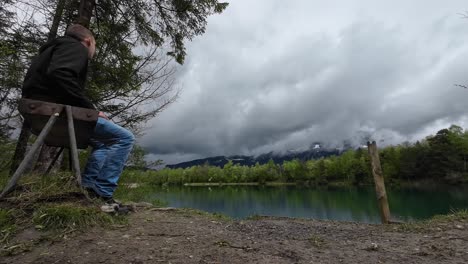 Guy-is-sitting-on-Bench-next-to-Mystical-Lake-with-turquoise-Water-on-a-Cloudy-Rainy-Day-in-Austria,-Europe