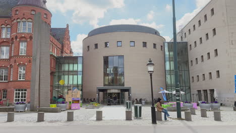 The-entrance-of-Malmo-Library-captures-urban-life-with-people-sitting-and-walking,-offering-a-glimpse-of-the-old-part-of-the-library