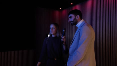 a-couple-of-young-people,-a-pretty-little-blonde-woman-and-a-embarrassed-dark-haired-man-with-a-beard-singing-in-a-karaoke-machine-with-a-microphone,-wooden-wall-with-coloured-lights-in-the-background