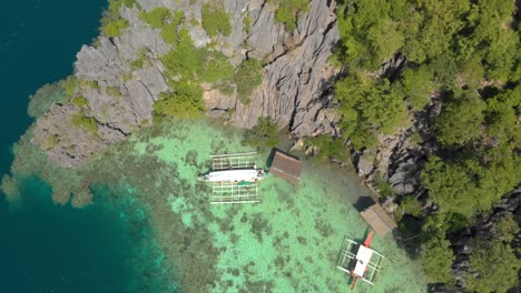 Drone-descending-toward-a-boat-with-outriggers-ties-up-at-a-small-hut-on-stilts-over-clear-blue-water