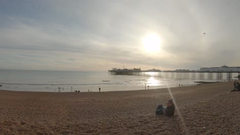 Gente-En-La-Playa-Con-Una-Puesta-De-Sol-Dorada-Y-Un-Destello-De-Sol-Sobre-El-Muelle-De-Brighton