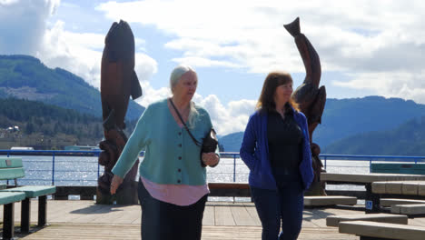 Senior-Mother-and-Daughter-Walking-Outdoor-Waterfront,-Sculptures-and-Mountains-in-Background