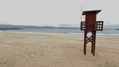 Cabaña-De-Madera-Elevada,-Puesto-De-Guardia-De-Los-Rescatistas-En-La-Playa-De-Arena-Fina-Frente-A-Los-Edificios-Y-Las-Montañas,-Día-De-Lluvia-Débil-Sin-Gente,-Panorámica-Descriptiva