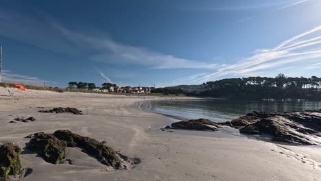 Una-Sombrilla-Vacía-Y-Una-Tumbona-En-La-Orilla-De-La-Playa,-La-Gente-Camina-Al-Amanecer-Con-Marea-Baja,-Cerca-De-Las-Casas-Unifamiliares-En-Verano-Un-Día-Soleado,-Tiro-Descriptivo-Bloqueado