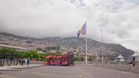 A-red-double-decker-bus-in-Funchal,-Madeira-with-a-mountainous-backdrop-and-cloudy-sky